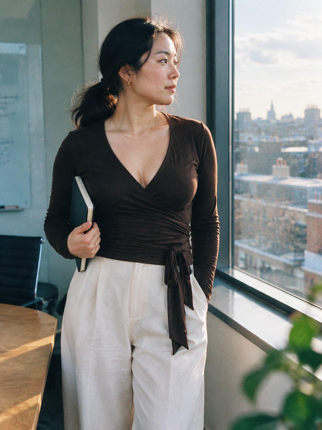 The Wrap Top in Brown, side view, standing by a window holding a notebook, styled with cream trousers