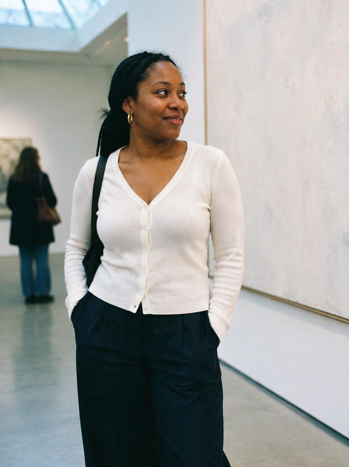 The Fine-Knit Cardigan in Cream, front view, walking through a gallery, styled with navy wide-leg trousers